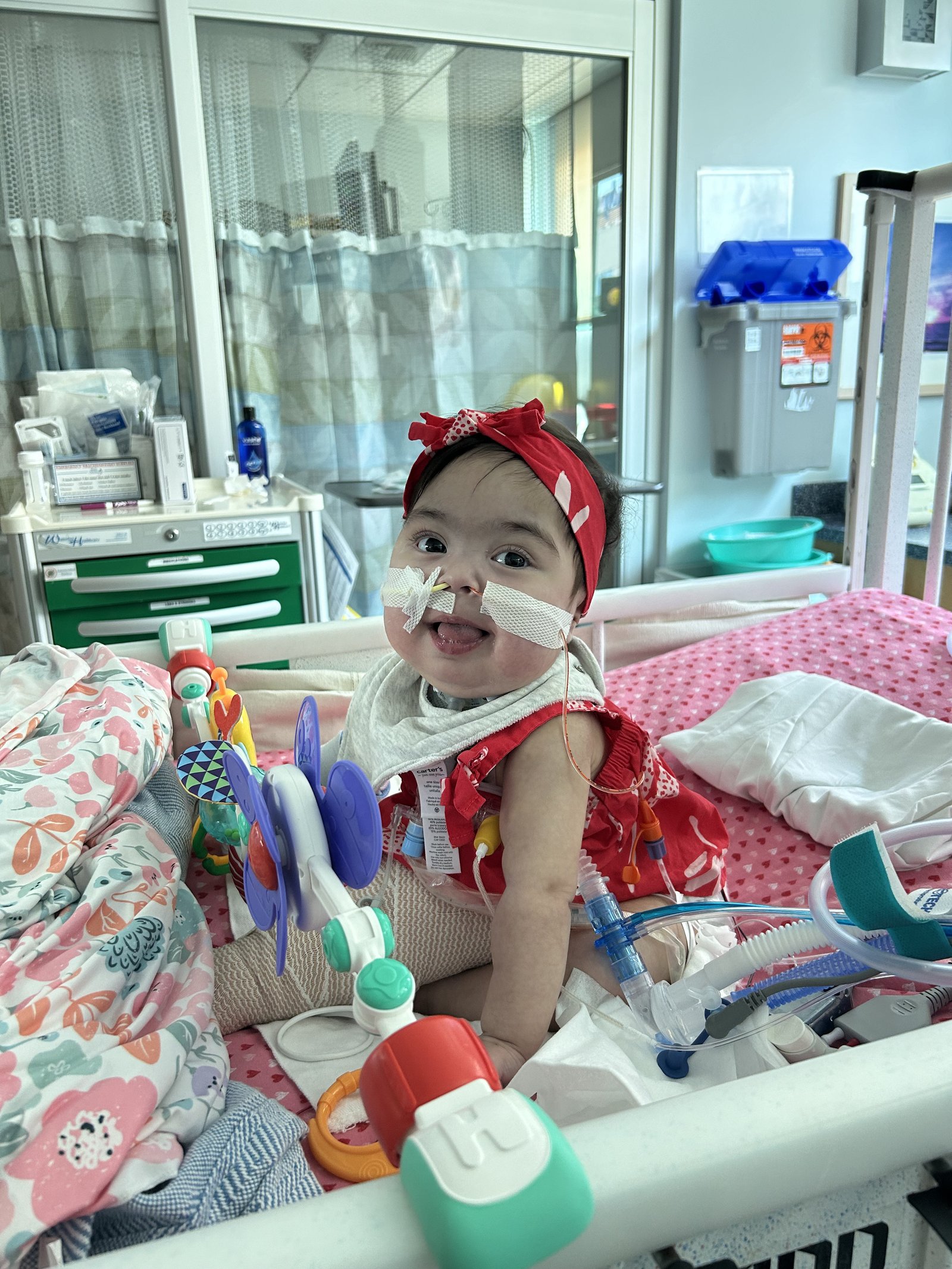 Baby Lynlee sitting up in her NICU bed wearing a red bow, smiling with her toys